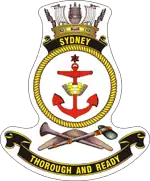 A ship's badge. A naval crown sits on top of a black scroll with "SYDNEY" written in gold. This is atop a yellow, rope-patterned ring, in which a red anchor is centred. Below the ring are a stone axe and a nulla nulla sitting on top of a boomerang. At the bottom of the badge is a black scroll with "THOROUGH AND READY" written.