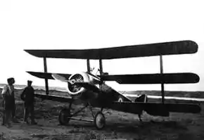 Front three-quarter view of military triplane on landing ground, with two men standing beside it