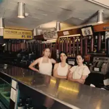 Este, Danielle, and Alana Haim dressed in white standing behind the counter of a deli. A sign with the album title is in the upper left corner.