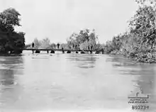 7 soldiers standing on pontoon bridge in middle distance; fast flowing w1ide River Jordan in foreground; lush growth on both banks.