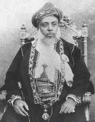 A black-and-white photograph of a man with a white beard wearing a turban, a dark jacket, a white shirt, and a belt and sitting on a chair