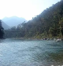 Hanging bridge over a river lined with tropical vegetation in a mountainous landscape.