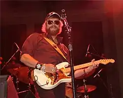 A bearded man wearing dark glasses and a cap, playing a guitar