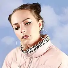 A brunette girl (Hannah Diamond) with hoop earrings, double buns, makeup, and a pink North Face jacket is looking down at the camera