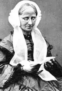 An older white woman seated, wearing a white bonnet with long wide ties; she has grey hair parted center, and is wearing glasses as she looks down into a small book.