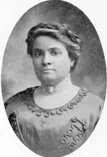 A woman with her hair in an updo, wearing a high-collared dress with a cameo at her throat; the outer laer of the dress is satiny, and embellished with heavy cord embroidery