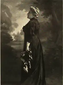Side view of standing woman with white hair in a bun wearing a long black dress with a high neckline