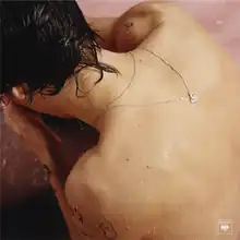 Harry Styles is half submerged in a pastel pink bath, both his hands cover his face while his back faces the viewer. Around his neck are two silver necklaces, with one of them flipped to his back to show a tiny lotus flower pendant.