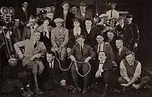 A black and white photo of the films cast gathered for a portrait, with a movie camera on the tripod on the left side