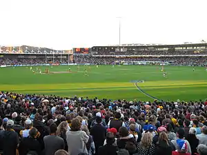 A small stand to the left and a two tier stand and scoreboard filled with people in the backdrop of an oval grass playing surface scattered with players. Spectators stand in the foreground.