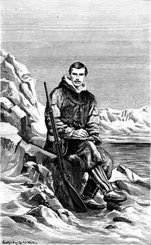 Heinrich Klutschak, in Inuit clothing, poses with a rifle, writing pad, and pencil in an arctic landscape