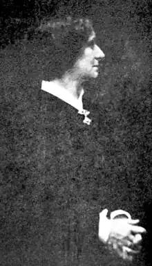 A white woman with dark hair, photographed in profile, wearing a dark dress with white collar and cuffs