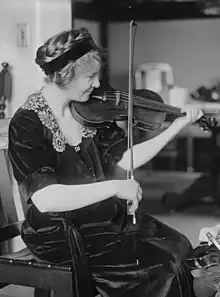 A young white woman seated, light hair braided across crown and held with a dark headband; she is smiling, and playing a violin; she is wearing a dark dress with a lace-trimmed neckline