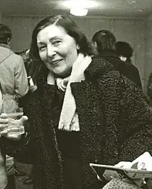 A woman in a heavy coat, holding a beverage in her right hand and a paper and a hat in her left hand.