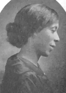 An African American woman, in profile. She is wearing a dark dress. Her hair is dressed into a chignon at the nape, and covers her ears.