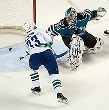 An ice hockey player wearing a white and blue jersey scores against a goaltender wearing a teal and black jersey with white pads. The player has both hands on his stick outstretched to direct the puck in the net, while the goaltender is off balance looking back at the puck in his net.