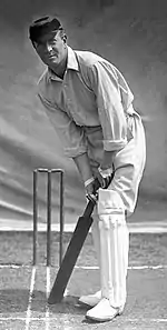 Man wearing white clothing, cap and leg pads positioned in front of stumps ready to bat the ball