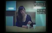 Old computer footage of a 37-year-old woman with long brown hair in a red jacket, sitting at a police interview desk. She is holding her hands together in front of her, and is looking to the right of the camera.