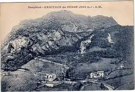 Old postcard with some buildings, including a castle at the foot of a mountain, with a road winding through vineyards.