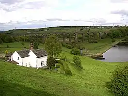 A white cottage in the foreground, a dam head and water in the middle ground, and a railway viaduct in the background