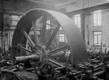 Flywheel for a large textile mill engine 1900, set up to machine grooves for the rope drives simultaneously. The saddle with two tool posts to the front. The wheel is rotated by two pinions driving via the cast-in barring gear teeth in the flywheel rim. Temporary wedges are securing the spokes to the hub of the wheel. A travelling crane behind and above.