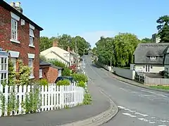 A red brick house in the foreground. Further down the street is a thatched house, behind which the church tower can just be seen, and across the road is a pub.