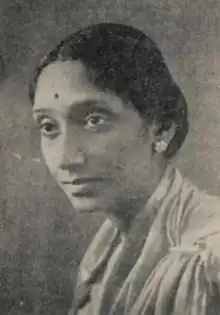 A South Asian woman, dark hair parted center and dressed to the nape; she is wearing a light-colored sari and earrings