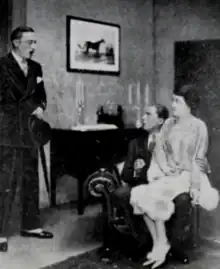 Smartly dressed young white man entering a room where a young man and women appear to be intimately seated