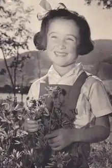 Black and white image of a young girl, in the 1930s, in a pastoral setting - behind her is a landscape of mountains and a lake, and before her a blooming wildflower