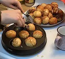 Homemade Danish æbleskiver, sometimes known as Danish pancakes, being removed using a fork from a cast-iron pan on the stove and set on a plate.