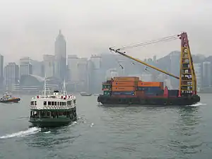 A lighter carrying shipping containers (loaded and unloaded by the tall derrick-crane on the lighter's top-deck) in Hong Kong's Victoria Harbour.