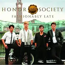 The band is dressed in prep school attire, with a building and a black car behind them. The band's name, logo and album title is placed above them, colored in gold.