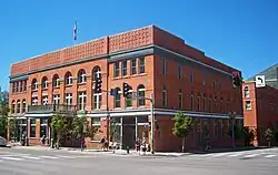 A three-story brick building, seen from its left front across a street with a traffic signal in front, and lit by the sun from its right. It has ornate windows, round-arched at the top, and a decorative wooden shelter at its main entrance. Atop the parapeted roof there is a flagpole.