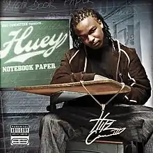 An image of a man wearing a brown hoodie, black t-shirt and faded blue jeans sitting on a classroom desk. Behind him is a chalkboard with the artist's logo and album title.