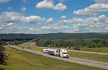 A divided expressway curves from the right of the image through a mostly wooded landscape towards a ridgeline at the rear below a blue sky filled with little white fluffy clouds. Along the roadway closest to the camera is a large white truck with "Perry's Ice Cream" along the side and pictures of scoops of ice cream in various colors and flavors