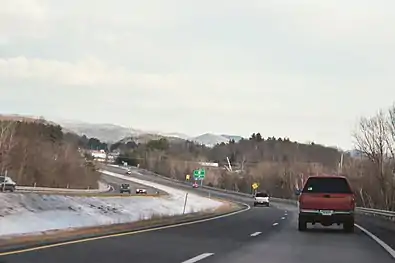 A four-lane highway in snowy weather, curving left with several cars on it. An exit sign and mountains are in the distance.