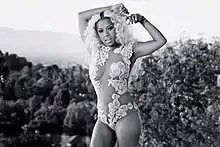 A black-and-white image of an African-American woman in a short sleeve one piece bikini posing in front of a pair of bushes on top of a hill.