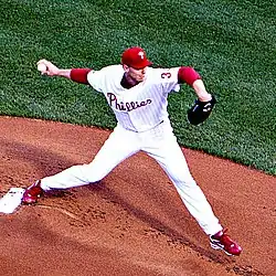 A man in a white baseball jersey and red baseball cap throws a baseball with his right hand. His jersey reads "Toronto" and "34" on the front in black block lettering outlined in blue, and he has a black baseball glove on his left hand.