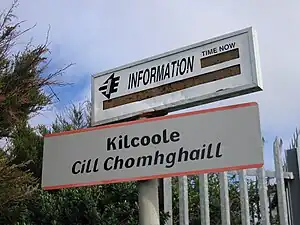 The name of the station i.e. Kilcoole, Cill Chomhghaill written in black font on white board with orange border on top and bottom. Another board with "Information" is written on white board above the name of the station. The board provides current time, and details of next train i.e. name and expected time of arrival. The details at the time of the picture are not clear. The picture is taken during the day.