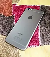 The Space Gray variant of an iPhone 6s showing its rear aluminum housing, identical to the iPhone 6. The "S" logo is the main visible difference from the iPhone 6.
