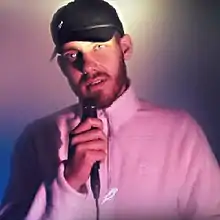 A bearded San Holo, wearing a black baseball hat and singing into a microphone