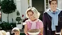  Image of a blond woman. She is a housewife and carries a pie in her left hand. Her hair is styled in 1950s fashion. She is wearing a pink polo shirt. Surrounding her are her husband and children.