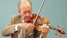 Ozim wearing a brown suit playing the violin against a blue background