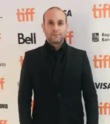 Ilan Eshkeri at the Toronto International Film Festival. He is standing against a white background with logos on it.  Eshkeri is wearing a black suit and has a neutral expression.