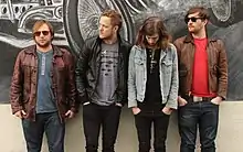 Four Caucasian men wearing jackets stand in front of a wall bearing a car mural.