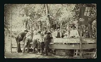 A group of men and young boys are standing at a stall set up in a park sometime between 1910 and 1925. Writing on the image indicates this is a park in Roche Perce.