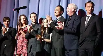 A man in a black suit, a woman in a pink dress, a man in a plaid suit, a woman in a black dress, a Japanese man in a black suit, and an old man in a blue suit clap their hands, while a man in a black suit stands. A microphone stand is in the foreground, and blue curtains are in the background.