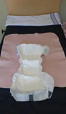 An adult diaper and a pink incontinence pad laid out on top of a single bed