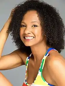 Indhira Serrano wearing a patterned sleeveless top, seated with elbow on one knee, head turned sideways, grinning at camera