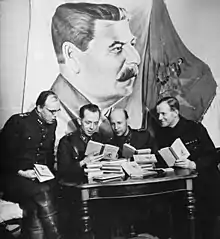 Four Finnish officers in uniform are sitting and reading Soviet skiing manuals with relaxed looks on their faces. A pile of the books is in front of them on a table, and a large drape of Joseph Stalin hangs above their heads on the wall.